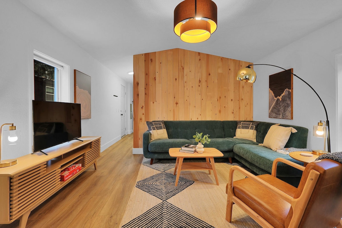 A mid-century modern living area features a green sectional sofa and wooden accent wall. A round coffee table sits in the center, complemented by a wood armchair. Natural light is allowed to flow through two nearby windows, enhancing the sophisticated design.