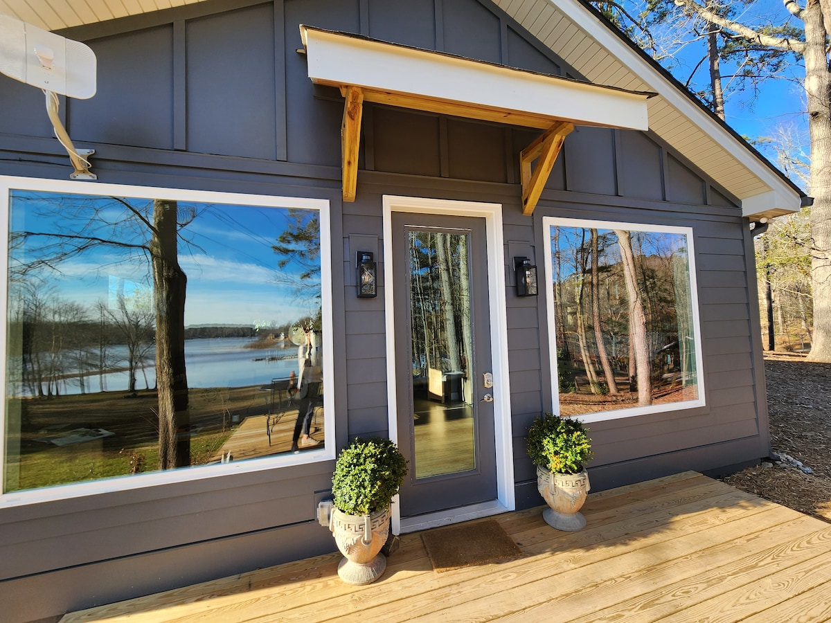 The exterior of the cabin features a modern design with large windows reflecting the serene lake and surrounding trees. A welcoming entrance is highlighted by a wooden porch with potted plants on either side, inviting guests to explore the tranquil setting.