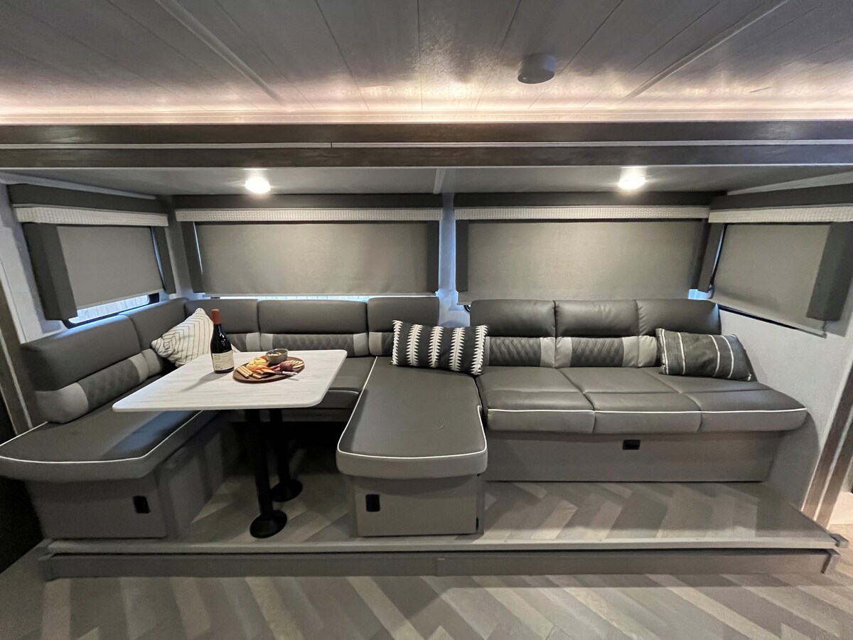 A modern seating area is presented with a comfortable sectional sofa featuring grey upholstery. A table is positioned at the center, ideal for dining or gatherings. Shades cover the windows, providing privacy and allowing adjustable light. Subtle lighting enhances the cozy ambiance.