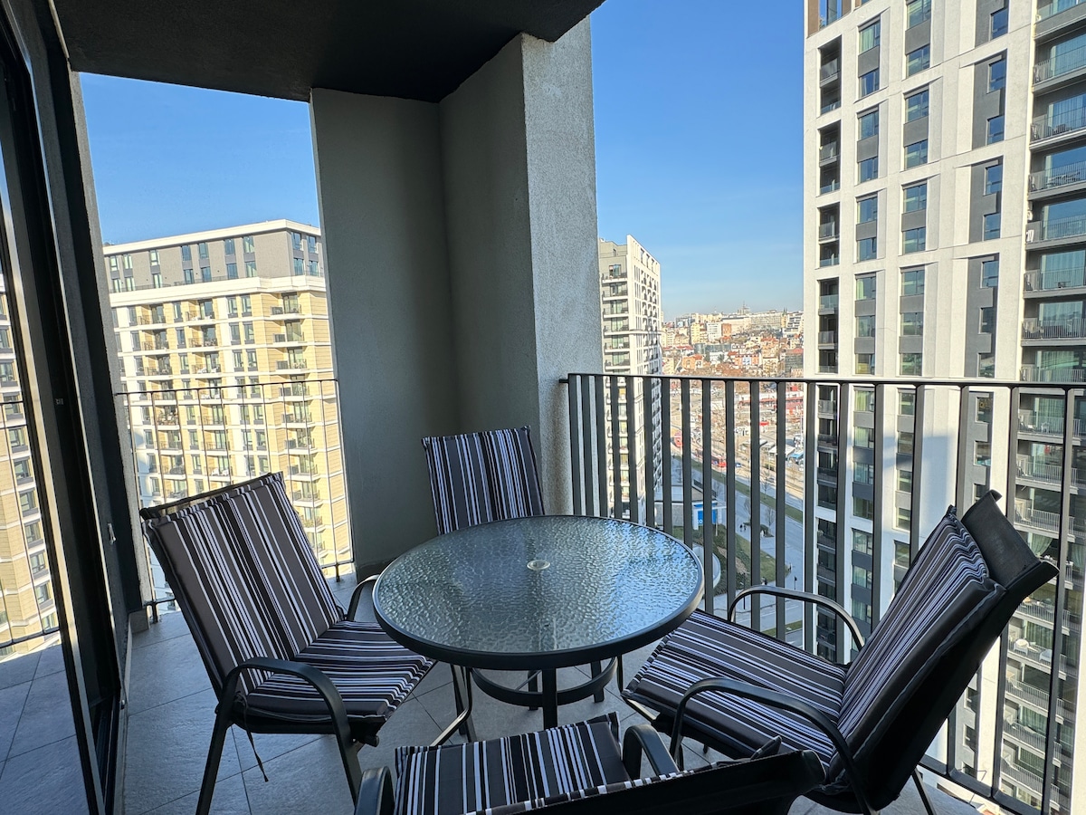 A balcony space is shown with a round glass-top table surrounded by four striped chairs. The city skyline is visible in the background, illuminated by natural light, emphasizing the modern architectural features of the neighboring buildings.