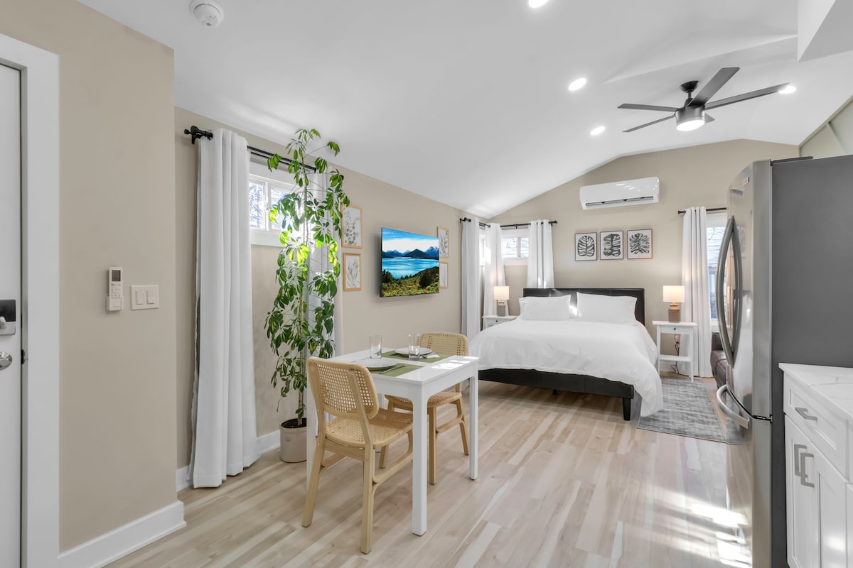 A studio apartment is highlighted by vaulted ceilings and a spacious layout. Natural light pours in through several windows, illuminating the plush queen-size bed. A dining table with two chairs is set beside a potted plant, and a kitchenette with modern appliances is partially visible.