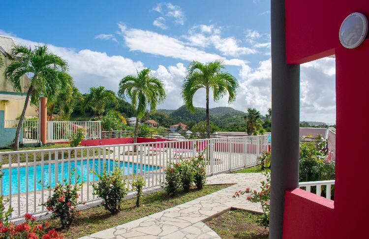 A pathway is framed by flowering shrubs, leading to a turquoise pool surrounded by palm trees. A vibrant red wall is visible in the foreground, while distant hills and more colorful buildings can be seen in the background under a clear blue sky.