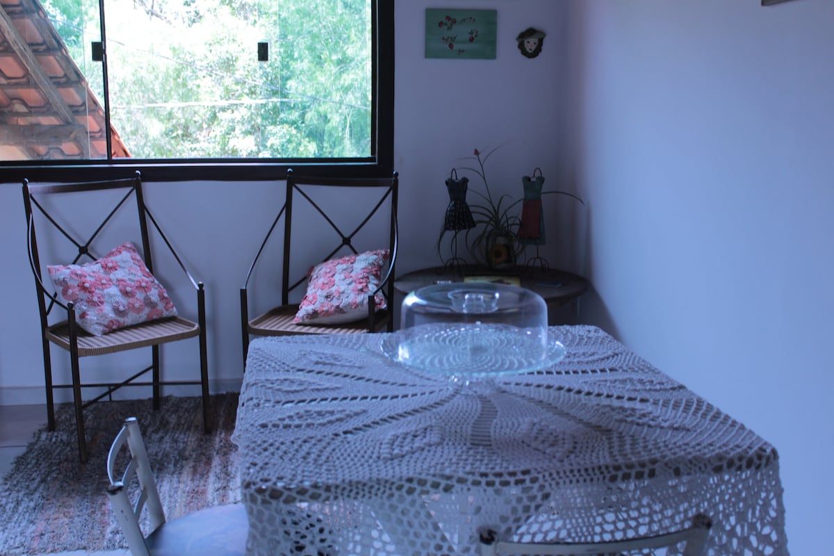 A dining area features a table with a lace tablecloth, surrounded by two chairs adorned with floral cushion covers. A decorative piece sits at the center of the table. Large windows allow natural light to illuminate the space, with greenery visible outside.