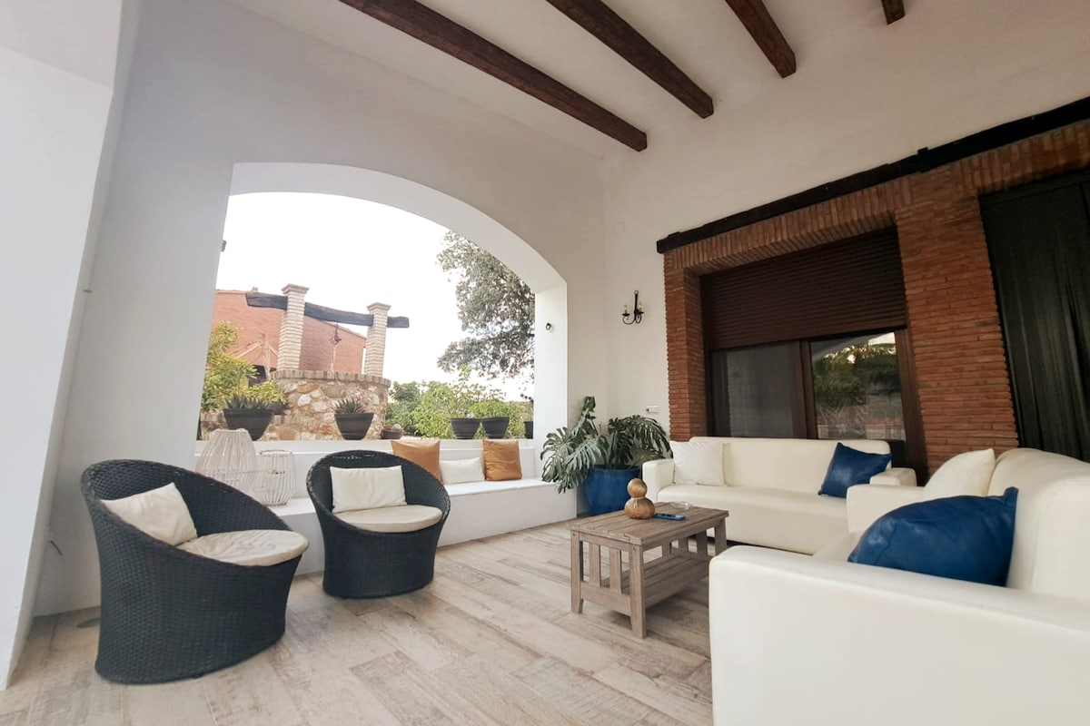 A spacious outdoor seating area is featured, containing a white sofa and armchairs with dark wicker frames. A wooden coffee table sits in the center. The area is enhanced by large windows allowing natural light and a view of the surrounding greenery.