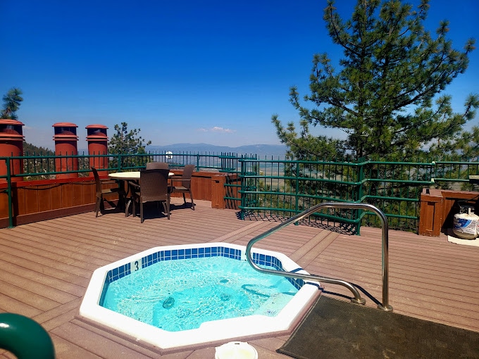A private hot tub sits on a spacious deck surrounded by greenery, offering mountain views. A patio dining set with chairs is nearby, complemented by a railing that enhances safety and aesthetic appeal. Clear skies provide ample natural light to the area.