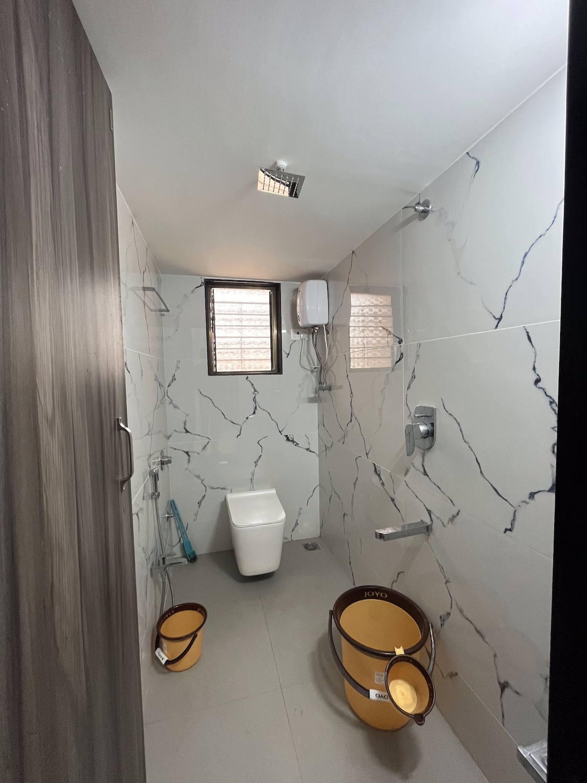 The bathroom features modern tiling with a marble design, including a shower area and a toilet. Two yellow waste bins are positioned on the floor, and natural light filters in through small windows. A wall-mounted shower and various fixtures are visible.