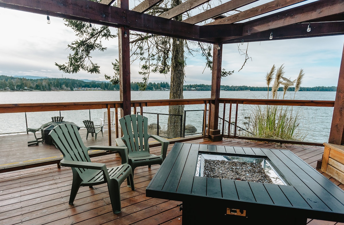 A spacious outdoor deck provides a tranquil space for relaxation, featuring five adirondack chairs arranged around a central fire pit. A scenic view of the water is visible, complemented by surrounding greenery and tall grasses swaying gently in the breeze.