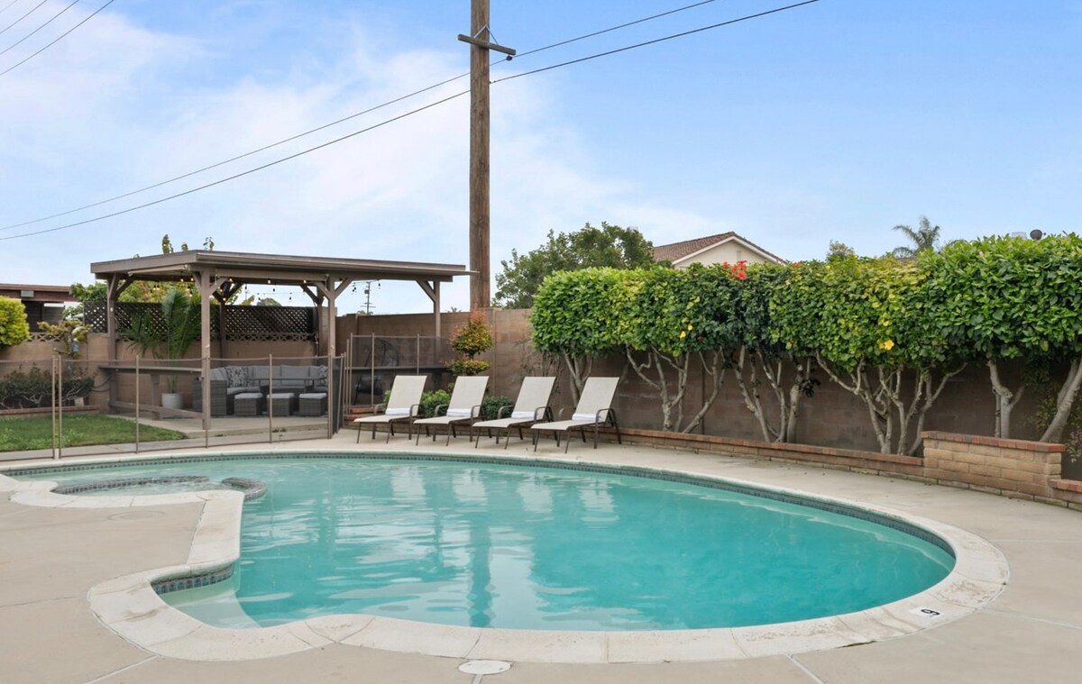 The outdoor area features a private pool surrounded by a spacious patio. Five loungers are positioned for sunbathing. A covered seating area is visible in the background, providing a shaded space for relaxation. Lush shrubbery lines the yard, contributing to a sense of privacy.