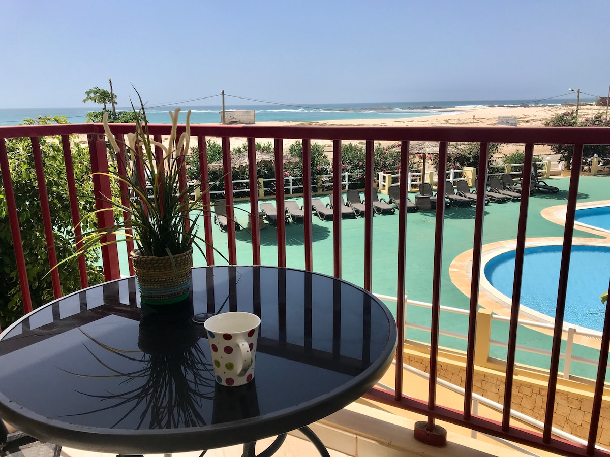 A small outdoor table is positioned on a balcony, featuring a potted plant and a cup. In the background, the swimming pool is visible, along with lounge chairs and expansive views of the ocean and sandy beach.