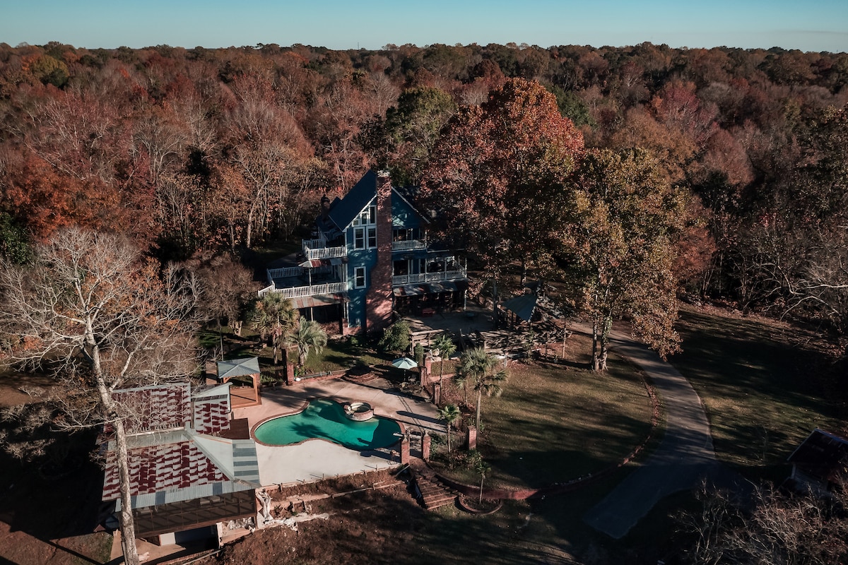 An aerial view showcases a spacious estate surrounded by vibrant autumn foliage. The home features multiple levels with large windows and balconies. A pool is visible, framed by a landscaped yard. A circular driveway leads towards the entrance, enhancing the property’s accessibility.