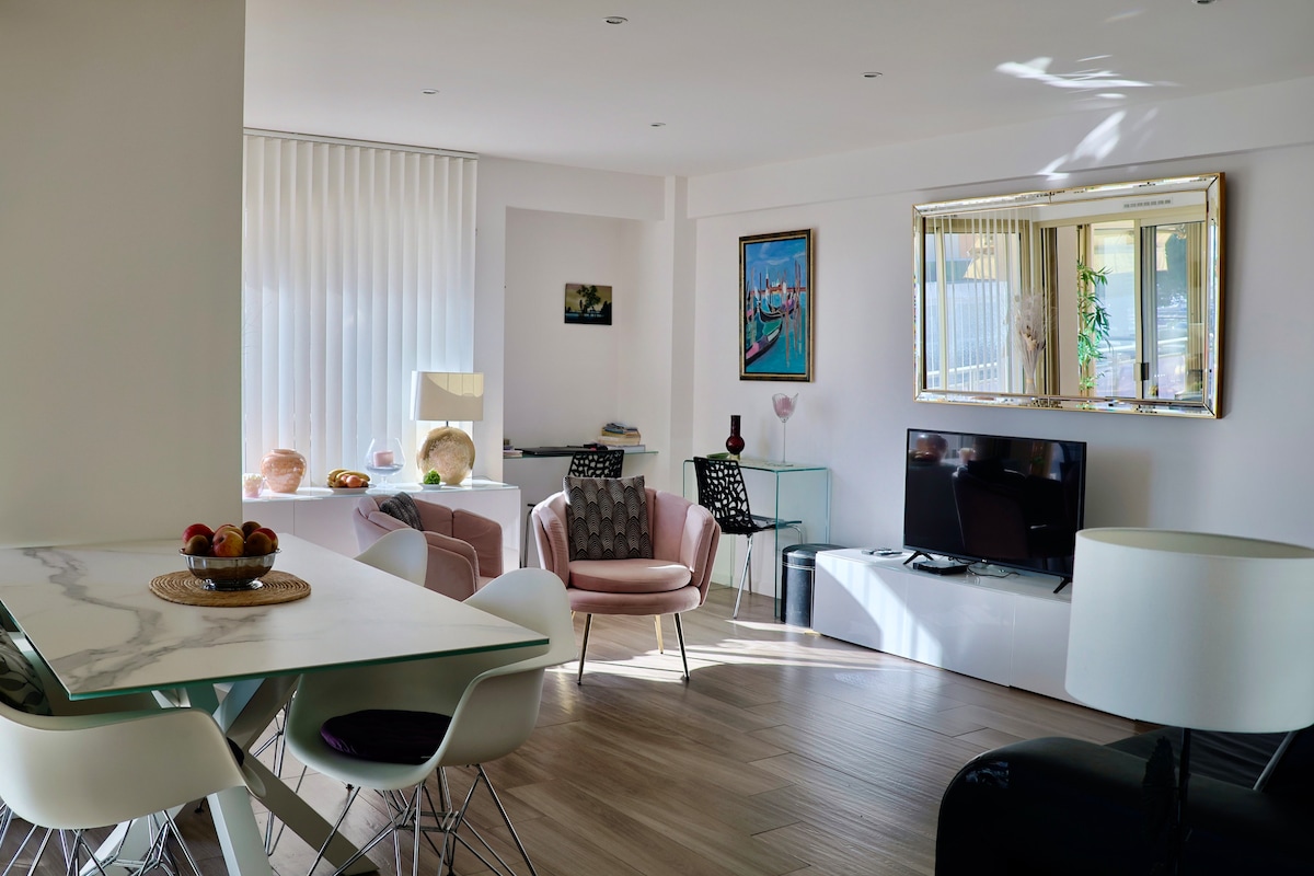 A bright living area features a dining table with decorative fruit, surrounded by modern chairs. A plush pink armchair and a sleek black sofa create inviting seating options. A large mirror reflects natural light, while a flat-screen TV is positioned on a low white cabinet.