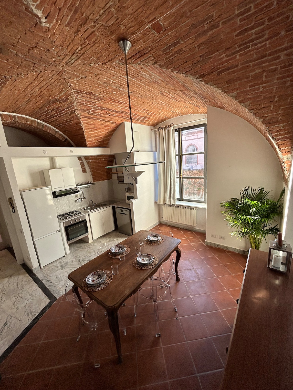 The kitchen area features high arched brick ceilings, creating a spacious ambiance. A wooden dining table is set for four, accompanied by clear chairs. Modern appliances are arranged along a marble counter, with a window providing natural light and a view of the outside greenery.