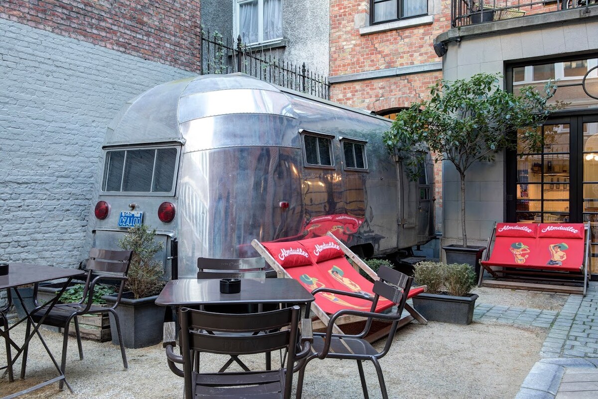 The exterior of a vintage Airstream Caravan is showcased, featuring its classic metallic finish. It is positioned in a courtyard surrounded by plants and outdoor seating, including wooden chairs and tables with brand decor. The environment reflects a blend of retro charm and modern outdoor comfort.