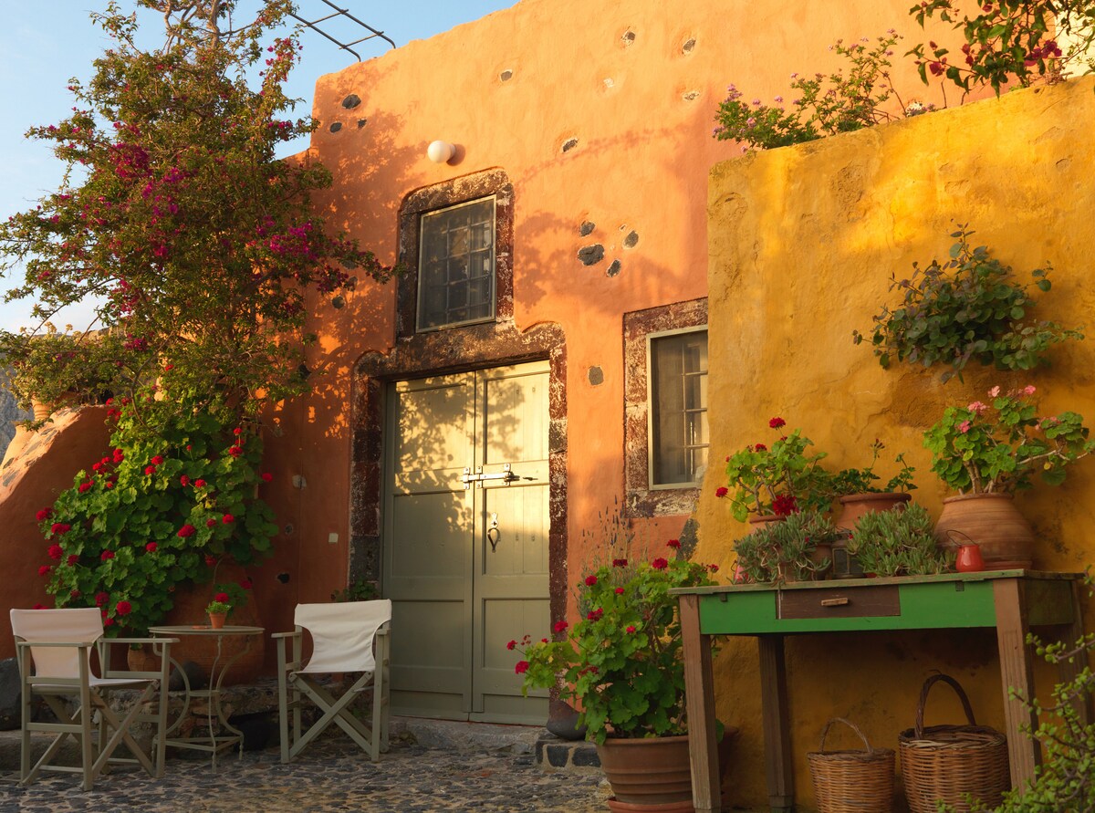 The exterior of the Pori House features warm, colorful walls adorned with blooming flowers. A green table sits beside two chairs, creating a cozy outdoor seating area. The entrance is framed by vintage windows, enhancing the traditional charm of the setting.
