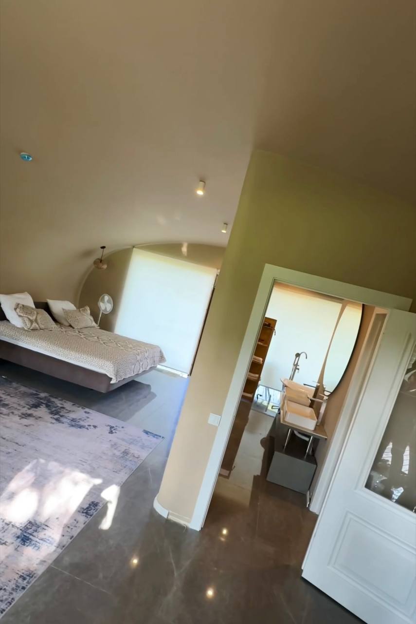 A spacious studio apartment features a large bed with neutral-toned bedding, illuminated by soft ceiling lights. An adjacent bathroom is visible, showcasing a shower area and modern fixtures. Natural light filters in through the window, creating a warm ambiance in the room.