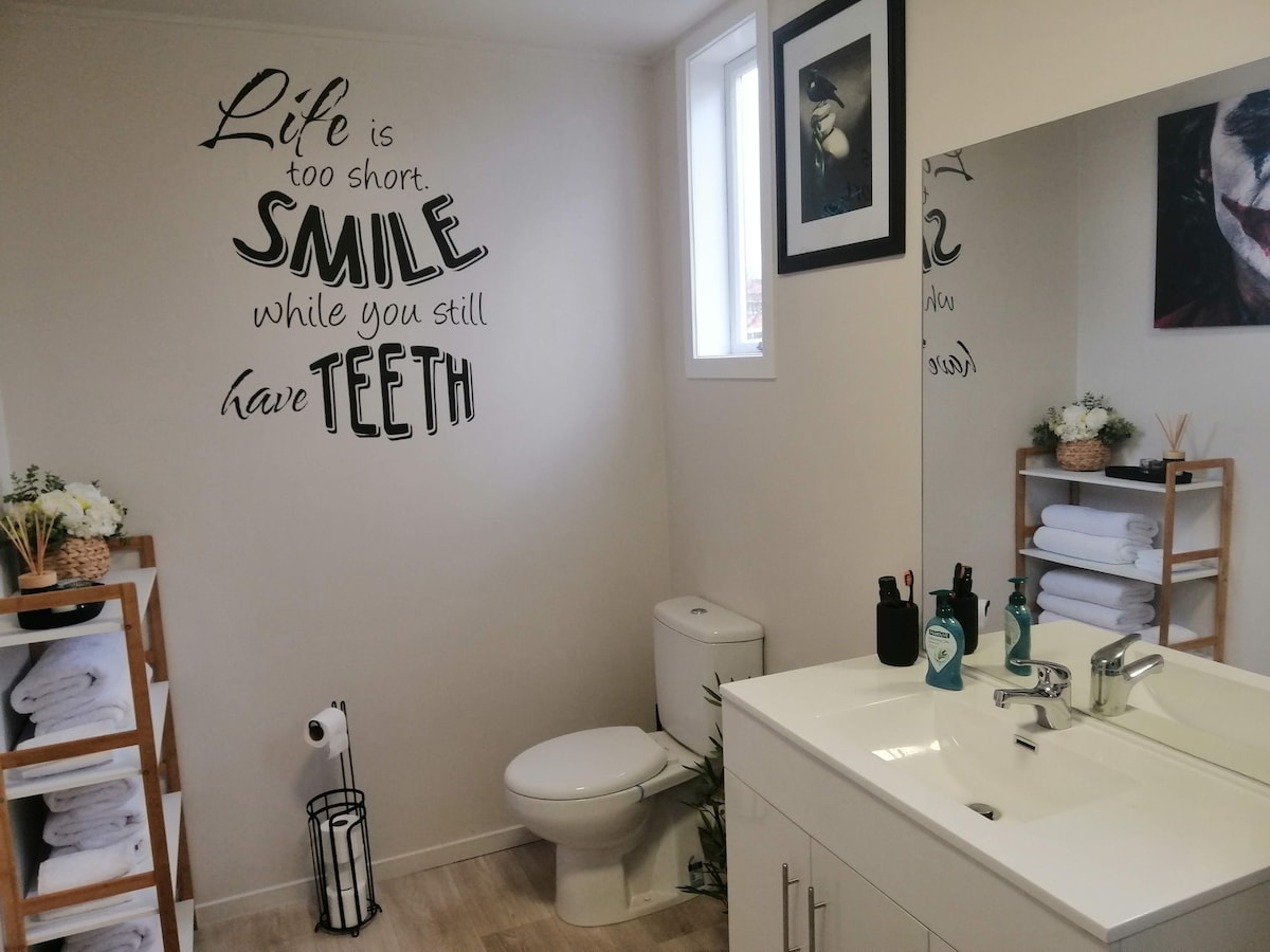 A modern bathroom features a clean, bright aesthetic with white cabinetry and a large mirror. A quote on the wall adds a playful touch. Fresh towels are neatly stored on a wooden rack, while greenery enhances the inviting ambiance.