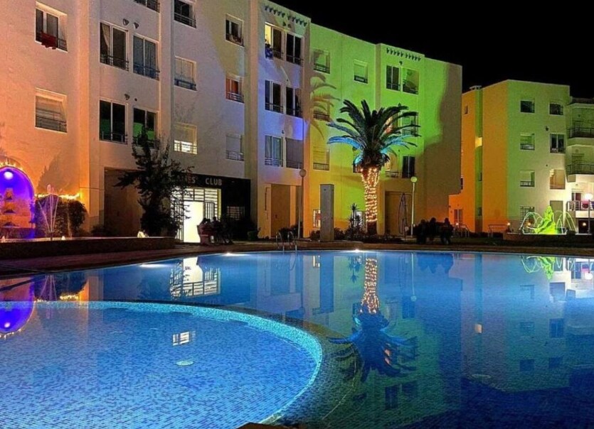 The nighttime view of the pool area showcases sparkling waters reflecting colorful illumination from the surrounding buildings. Palm trees frame the scene, enhancing the tranquil atmosphere of the landscaped outdoor space.