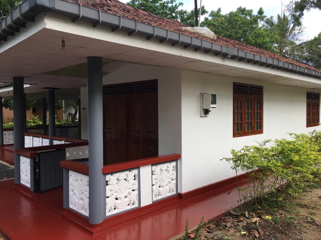 The exterior of the home features a traditional design with a red-tiled roof and intricate white decorative paneling. Multiple windows provide natural light, and a welcoming porch is outlined by polished railings, surrounded by lush greenery.
