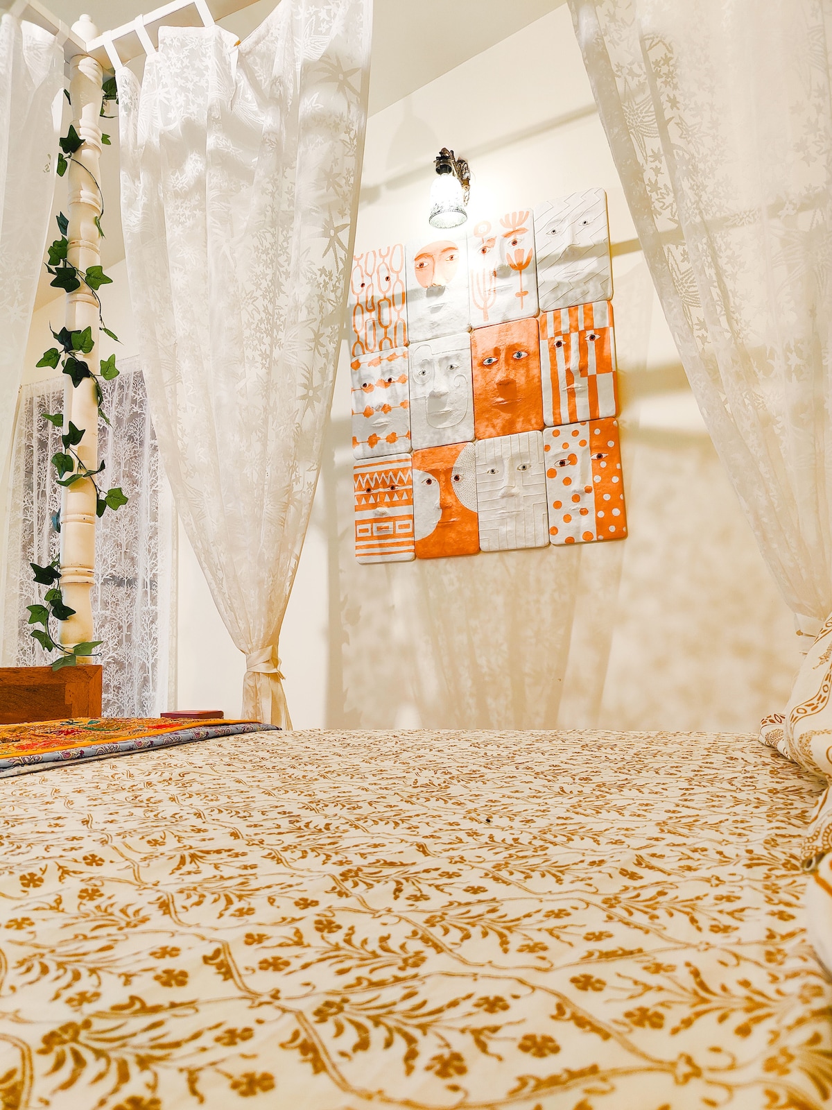 A spacious bed is adorned with a detailed floral coverlet, complemented by sheer, decorative curtains that frame the space. A vibrant wall display featuring an array of colorful art pieces adds character, while soft lighting casts a gentle glow throughout the room.