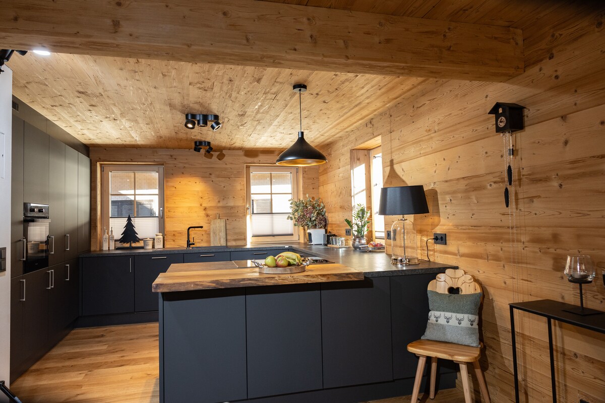 An open kitchen displays a central cooking island with a wooden countertop. Modern cabinetry features a minimalist design, while pendant lights illuminate the space. Windows allow natural light to enhance the warm wooden walls, complemented by decorative elements like a potted plant and a small chair.