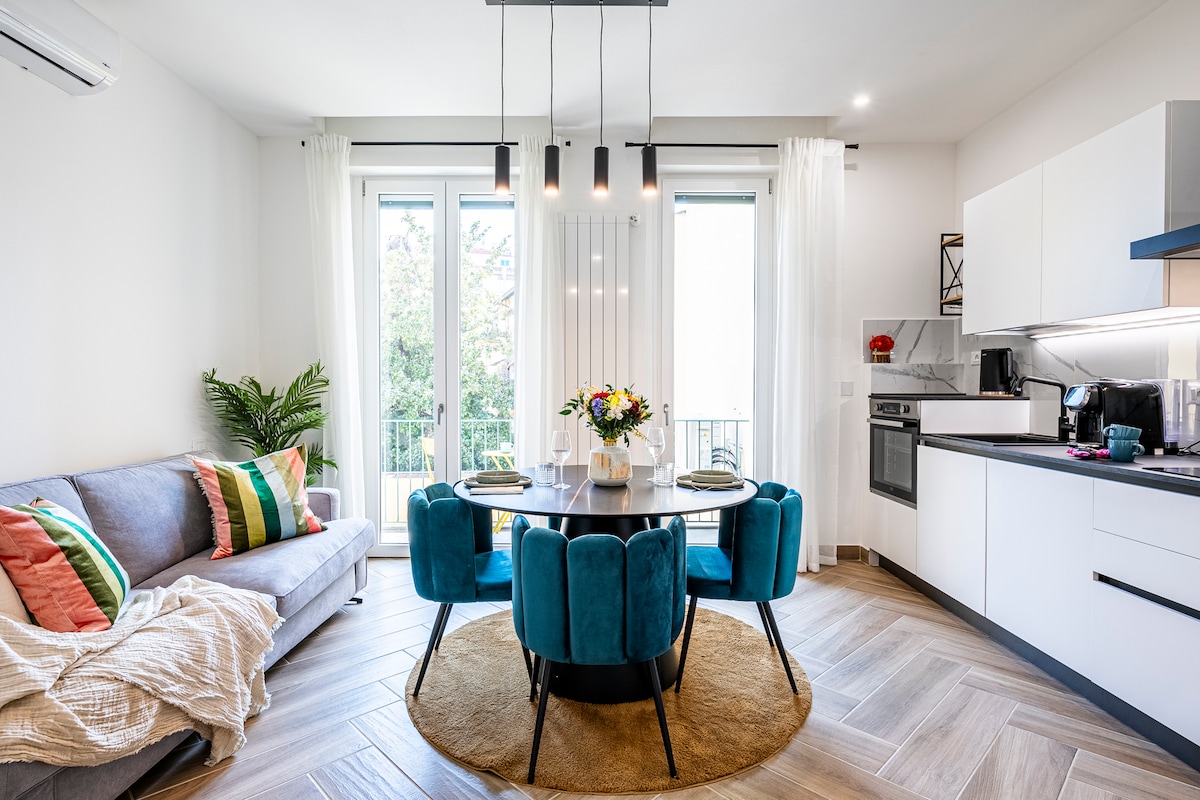 A modern living area features bright natural light streaming through two large windows, framed by sheer white curtains. A comfortable gray sofa, accented with colorful cushions, complements a circular dining table surrounded by plush blue chairs. A decorative vase filled with fresh flowers centers the table.