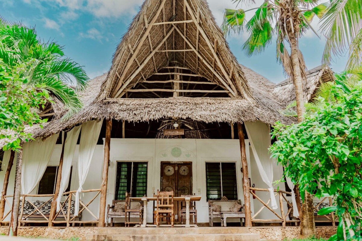The exterior features a thatched roof structure with open sides, surrounded by lush greenery. A wooden dining table is positioned on a patio adorned with comfortable seating. Light drapes flutter gently in the breeze, enhancing the inviting atmosphere of this outdoor space.