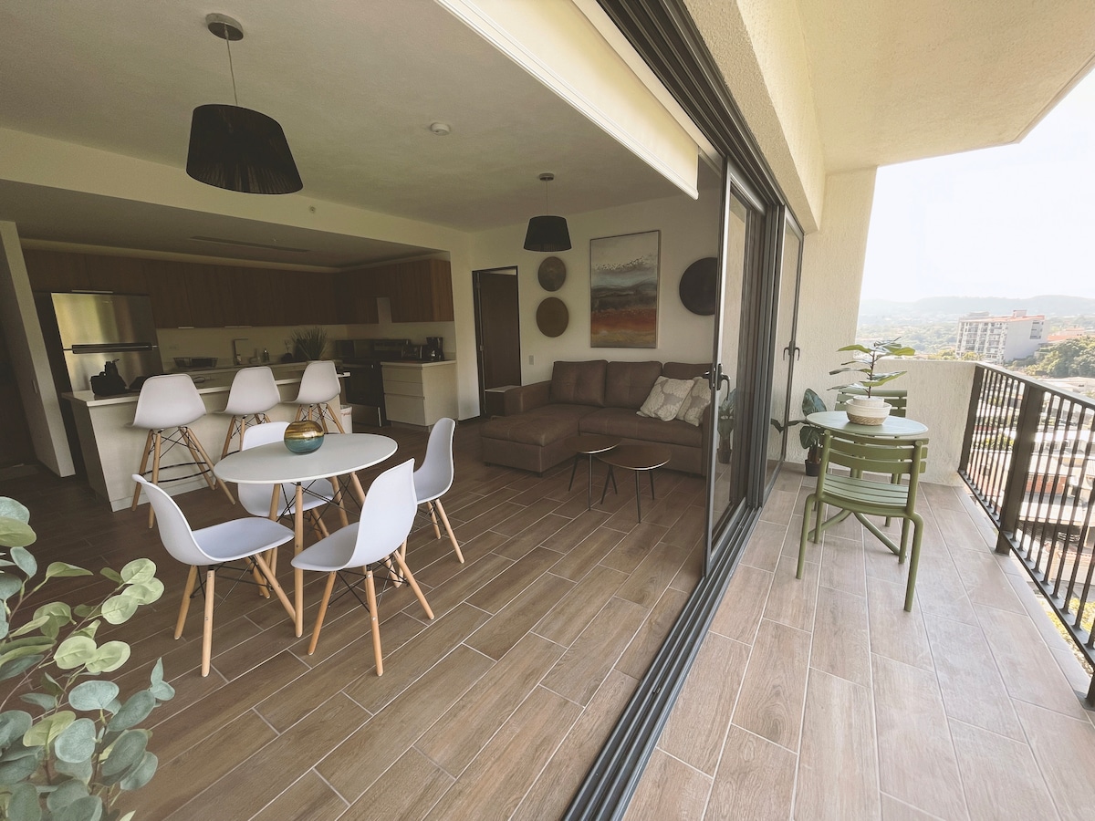 A modern open-plan living area is displayed, featuring a light-colored dining table surrounded by white chairs. A cozy seating area with a brown sectional couch is visible. Large glass doors open to a balcony with green chairs, offering panoramic views of the surrounding landscape.