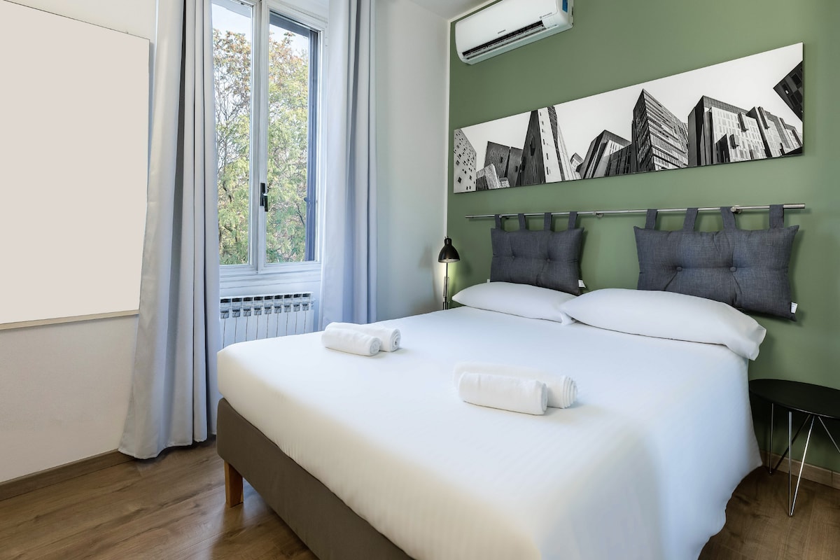 A bedroom features a spacious bed with white linens and two rolled towels neatly placed at the foot. A wall-mounted artwork displays a monochromatic cityscape. Natural light enters through a window adorned with light curtains, and an air conditioning unit is installed above.