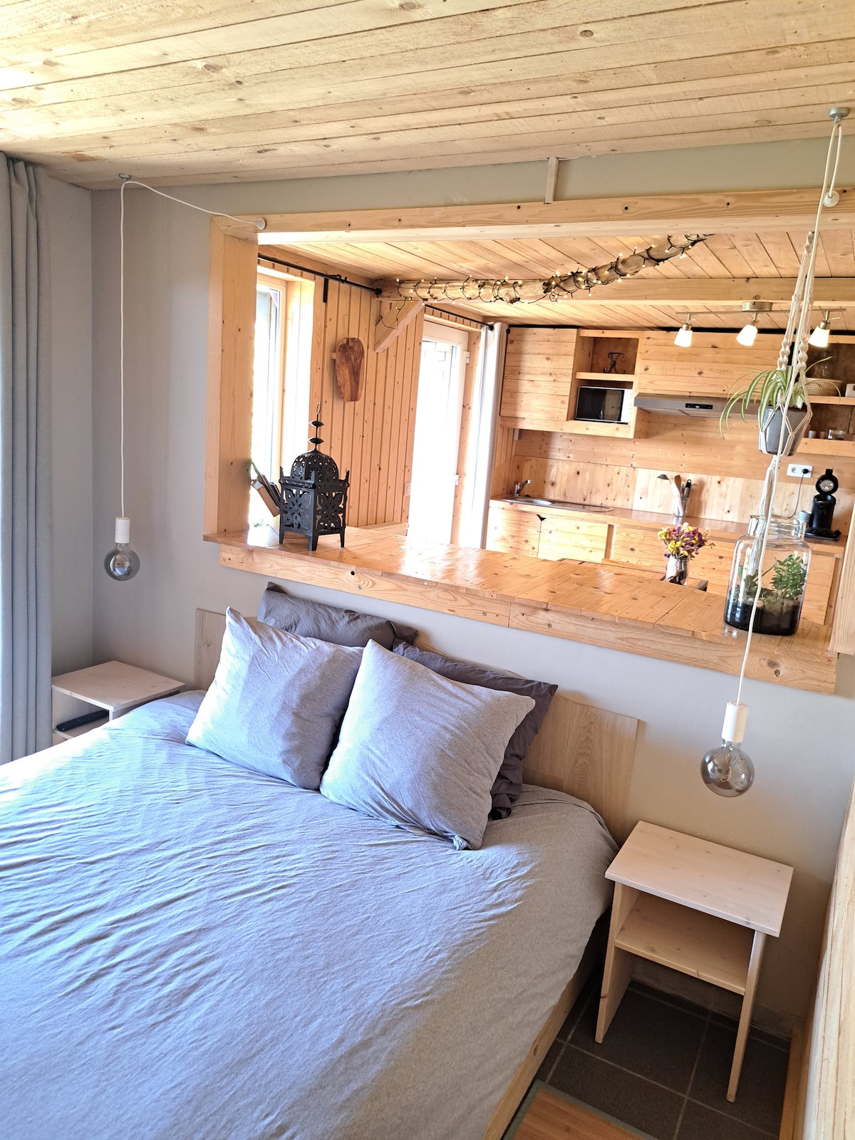 A serene sleeping area is highlighted by a spacious bed with gray bedding and two bedside tables. Soft pendant lights are mounted above each table. A view of the kitchen area is visible through a wooden pass-through, featuring natural wood accents.