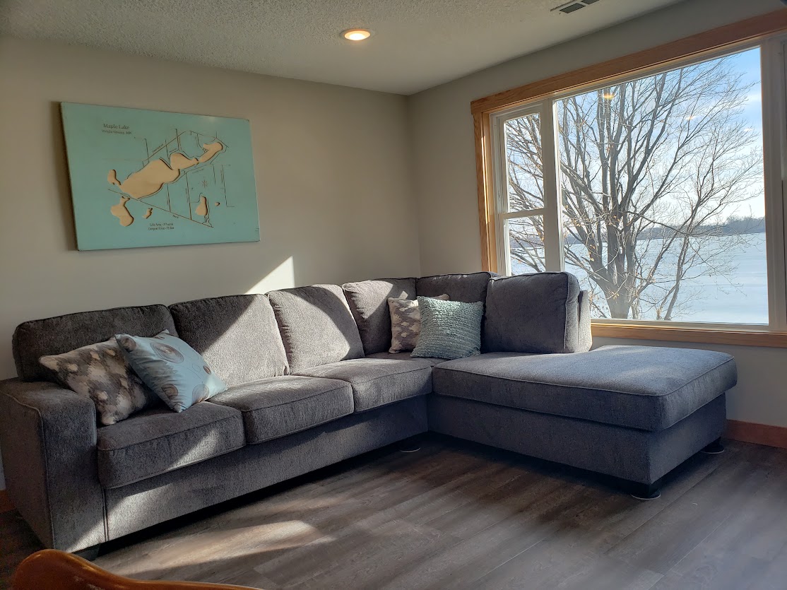 A cozy living area features a grey sectional sofa adorned with decorative pillows. Natural light floods in through a large window, offering views of the serene lake and surrounding trees outside.