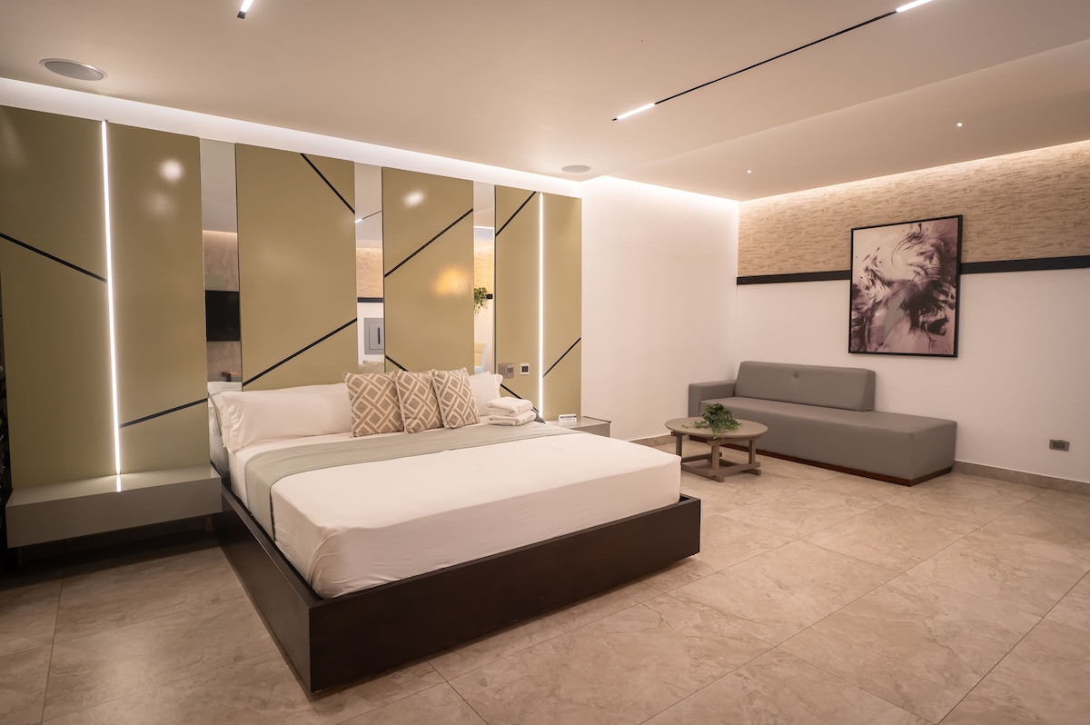 A spacious sleeping area features a large bed with neutral-toned bedding and decorative pillows. A modern gray sofa and a small coffee table complement the room, with geometric accent walls and soft lighting enhancing the contemporary design.