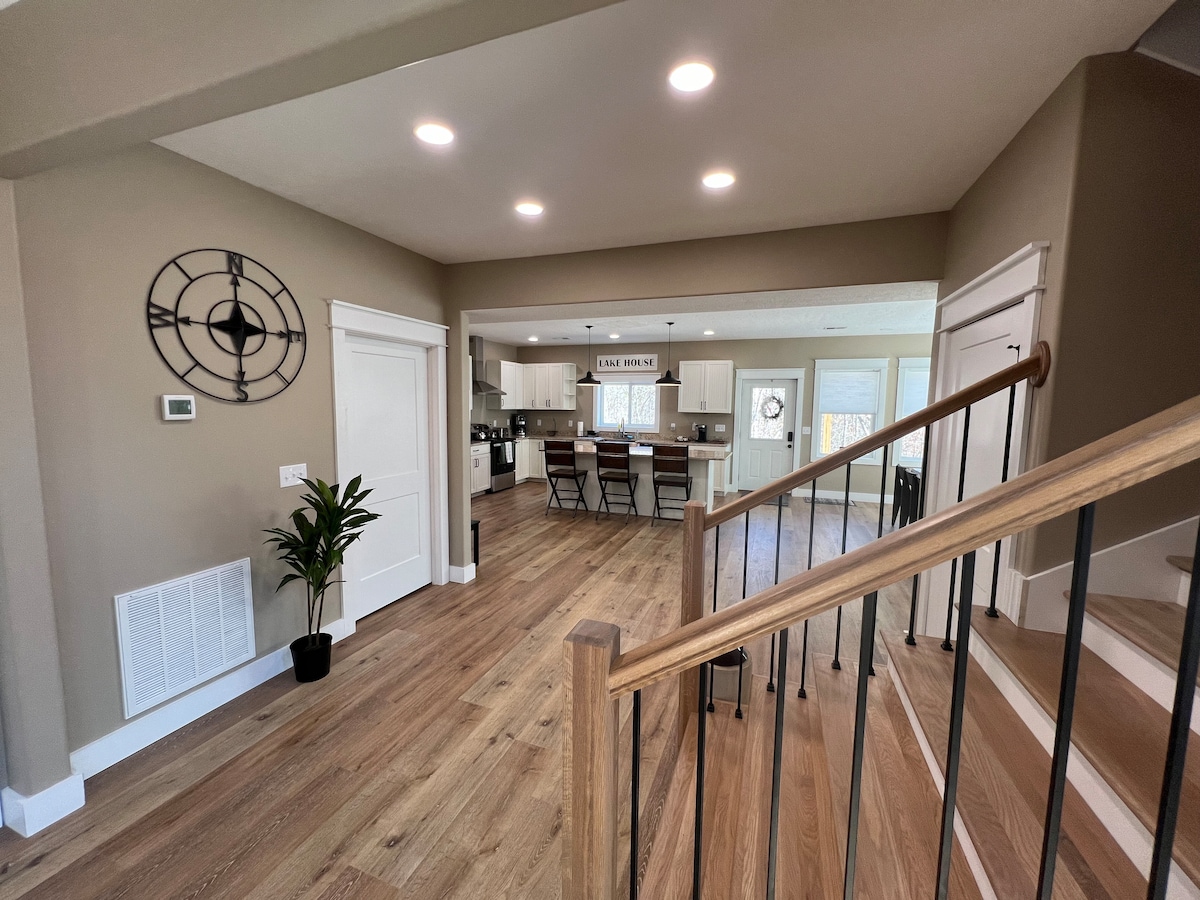 A spacious entryway features a wooden staircase leading to the second floor. The area is illuminated by recessed lighting and natural light, with a decorative wall clock and a potted plant enhancing the modern design. The open layout leads to a dining area and kitchen.