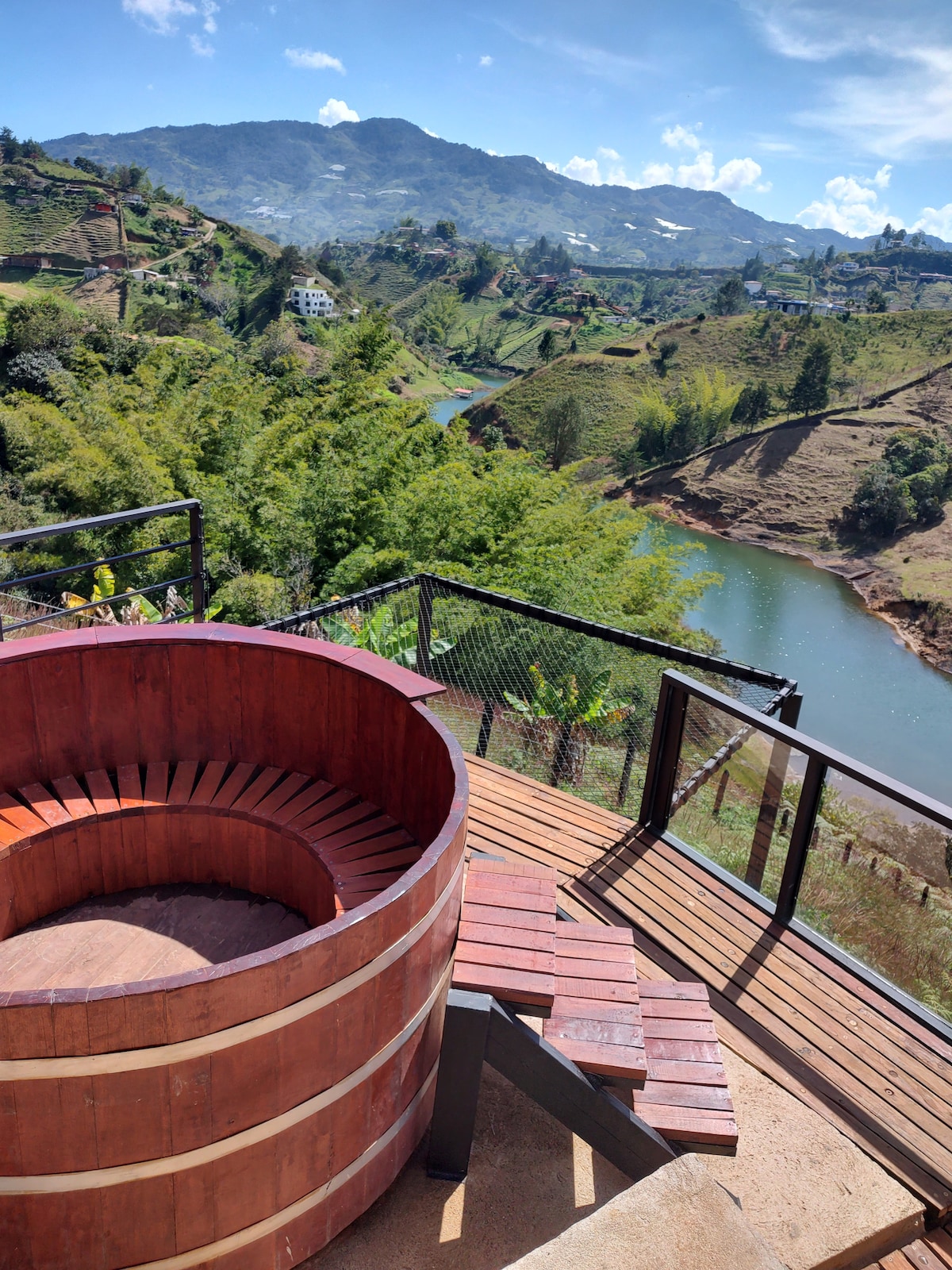 A wooden Japanese soaking tub, positioned on a wooden deck, overlooks a scenic landscape of hills and a river. Sunlight enhances the natural beauty of the surroundings, and the sky is dotted with clouds, creating a serene outdoor setting.