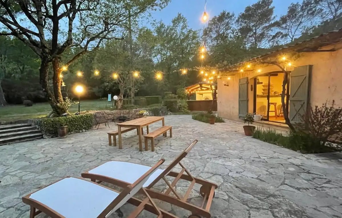 An outdoor area is highlighted with stone flooring and wooden furniture, including lounge chairs and a dining table. Warm lighting hangs overhead, creating a relaxed ambiance. The entrance to the charming house is visible, surrounded by greenery and well-tended landscaping.
