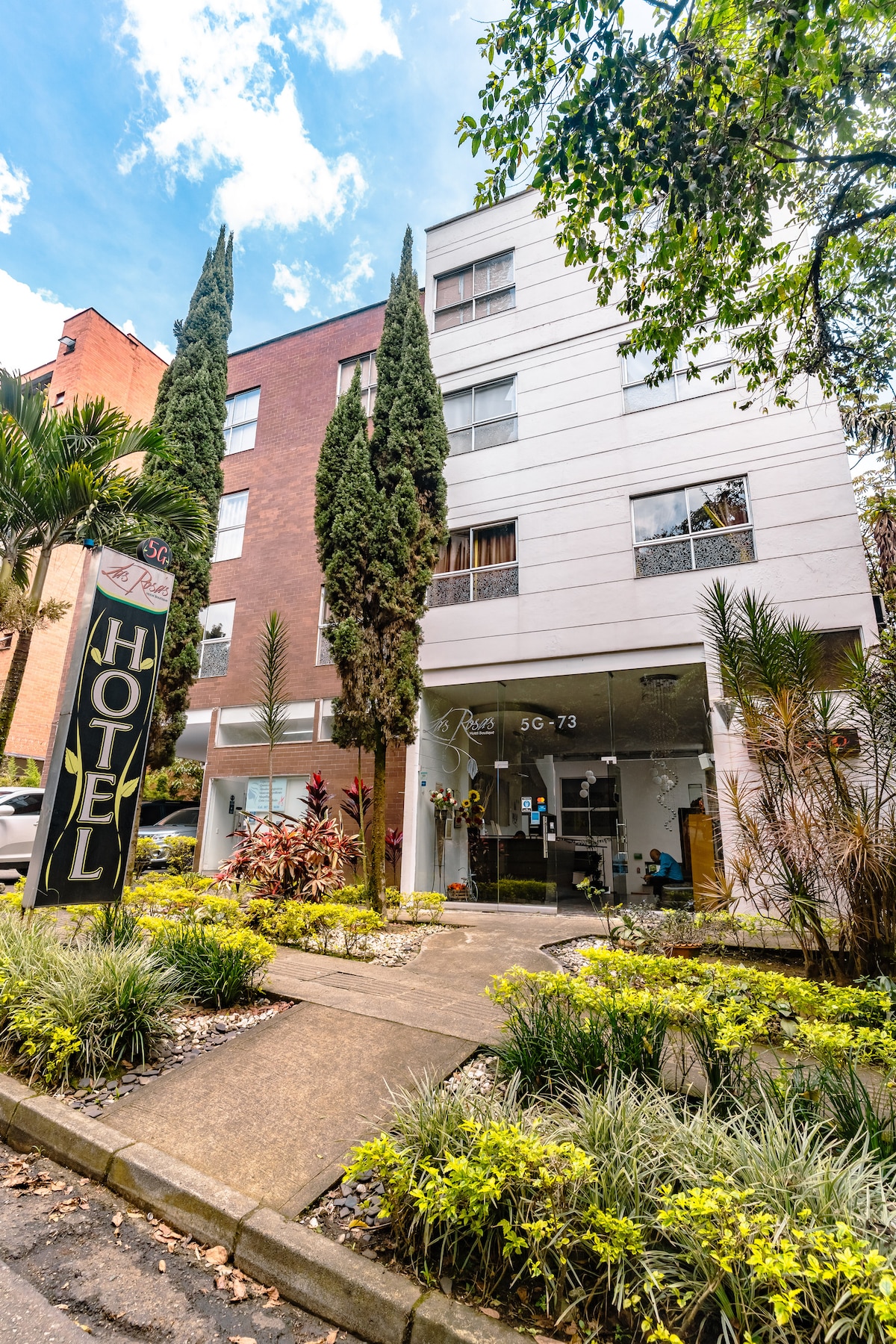 The exterior of the hotel is presented, featuring a modern three-story building with a blend of brick and light-colored siding. Lush greenery and decorative plants frame the entrance, with a prominent sign indicating 'HOTEL.' Clear skies are visible above, enhancing the welcoming atmosphere.