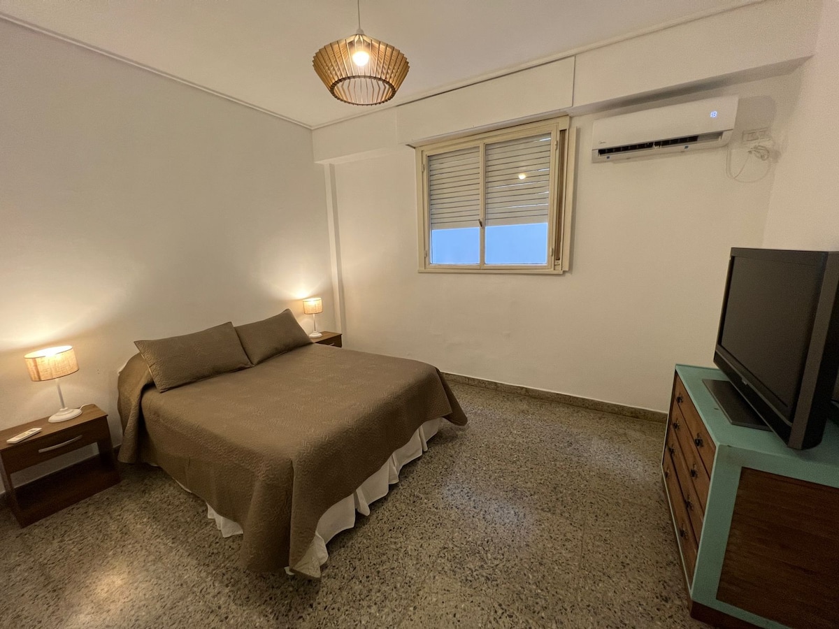 A well-lit sleeping area features a spacious bed with a neutral brown coverlet. Two bedside lamps provide soft illumination. A small television is positioned on a light blue cabinet, while a window allows natural light to enter, framed by simple white curtains.