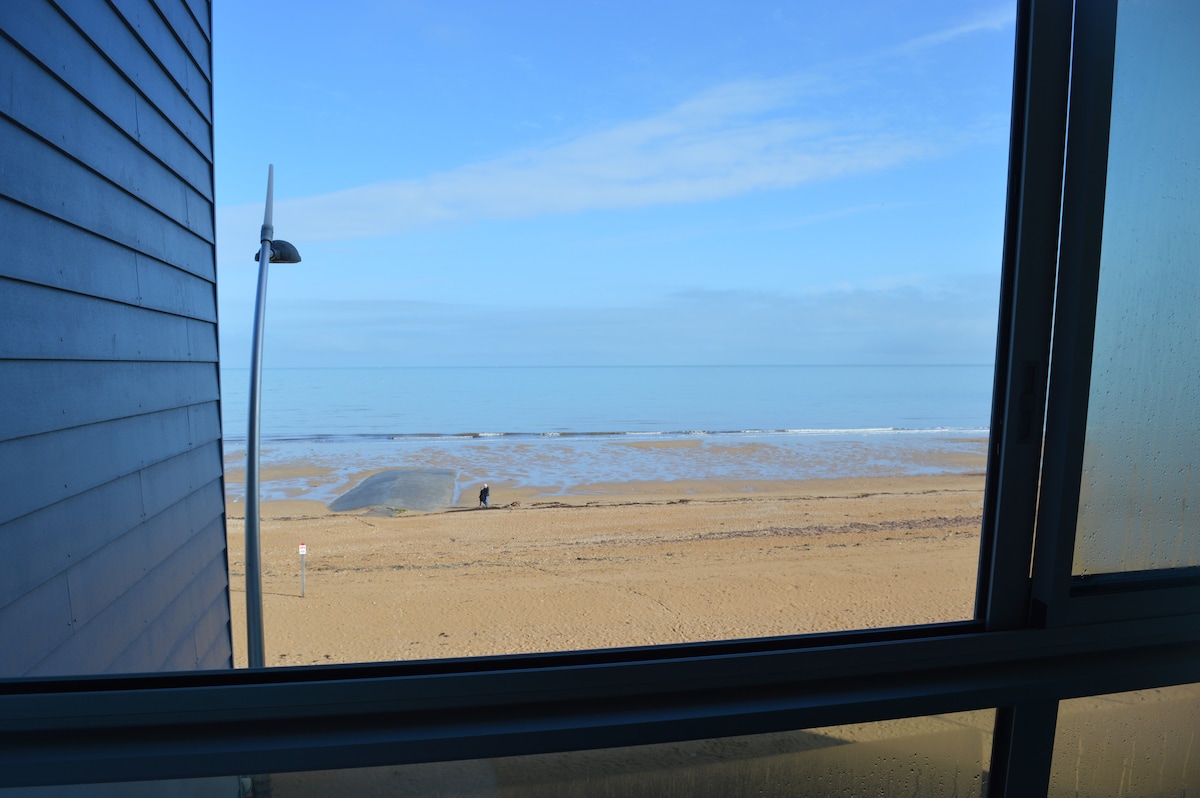 A view from the loggia showcases a sandy beach extending towards a calm sea. The horizon meets the sky, which is clear with soft blue tones. The scene is framed by the large window, enhancing the openness of the space.