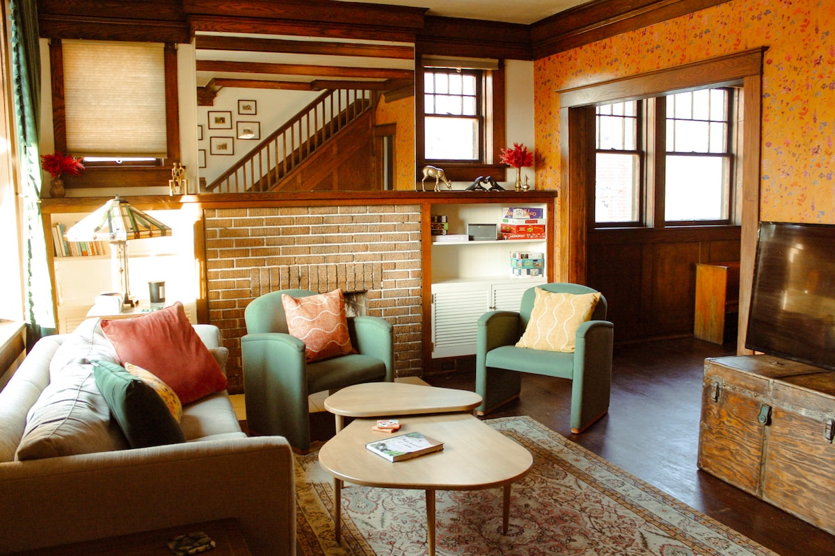 A cozy living area features a comfortable sofa and two armchairs arranged around a wooden coffee table. A patterned area rug lies on the dark hardwood floor. A brick fireplace serves as a focal point, while warm tones and floral wallpaper add character.