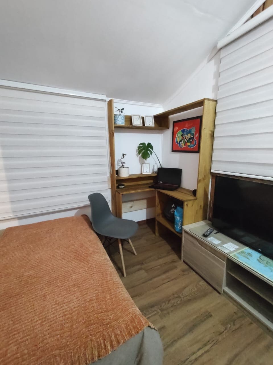 A functional corner workspace features a compact desk with a laptop and decorative items above. A comfortable grey chair sits nearby. The cozy bed with an orange throw is positioned near a wall-mounted TV, complemented by natural light filtered through window shades.