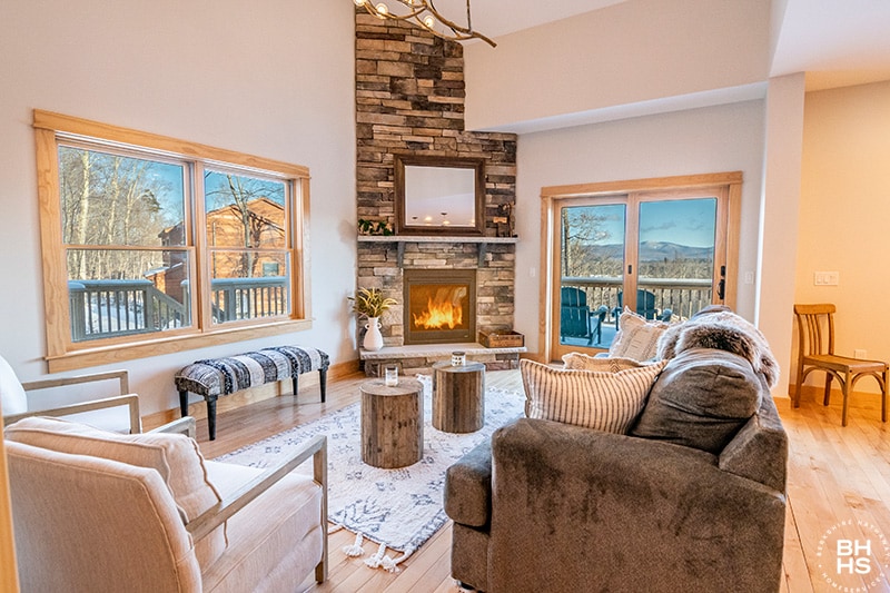A cozy living room is showcased with a stone fireplace as the focal point, surrounded by comfortable seating. Large windows frame mountain views and natural light floods the space, while a woven area rug adds texture to the hardwood floor.