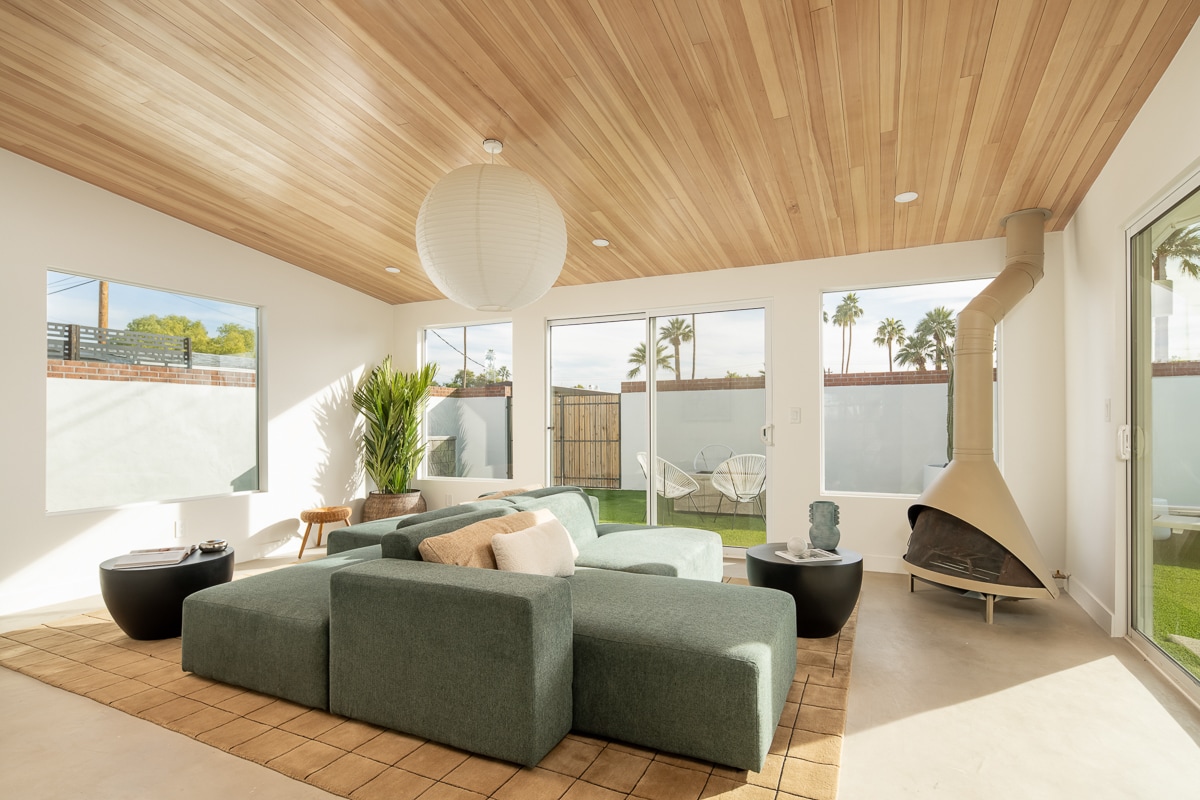 An open living area features a modern sectional sofa and two round coffee tables atop a neutral area rug. Sunlight enters through large glass doors, illuminating the wood-paneled ceiling and minimalist decor. A unique fireplace sits in the corner, enhancing the contemporary design.