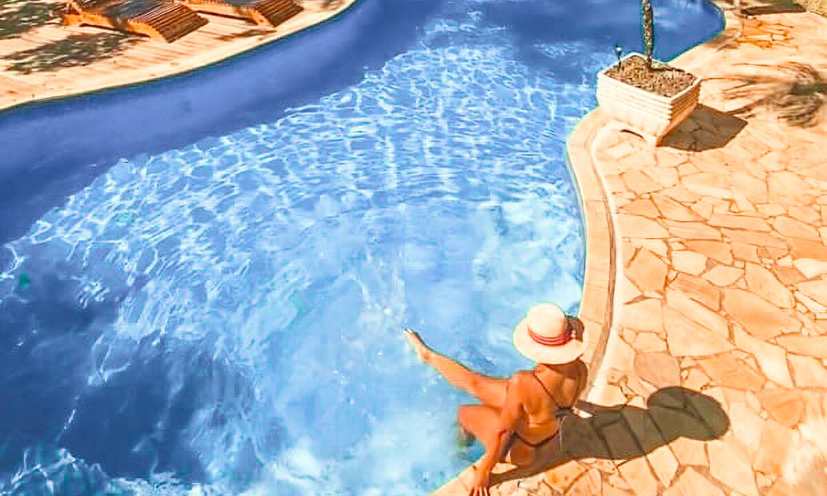 A curved pool with crystal-clear water is surrounded by a stone deck. A person is seated at the pool's edge, partially submerged, wearing a sun hat. Sun loungers are visible in the background, suggesting a leisure area.