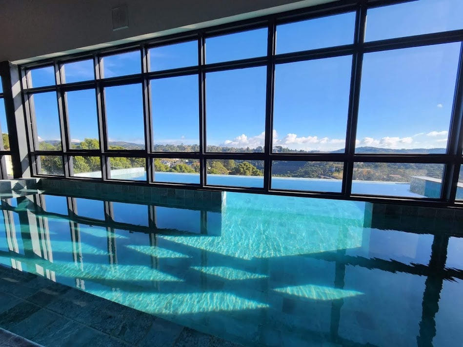 A serene indoor swimming pool is visible, featuring clear turquoise water that reflects sunlight. Large glass windows frame a view of lush greenery and distant mountain ridges, creating a bright and airy atmosphere. The pool appears to extend towards an outside infinity edge.