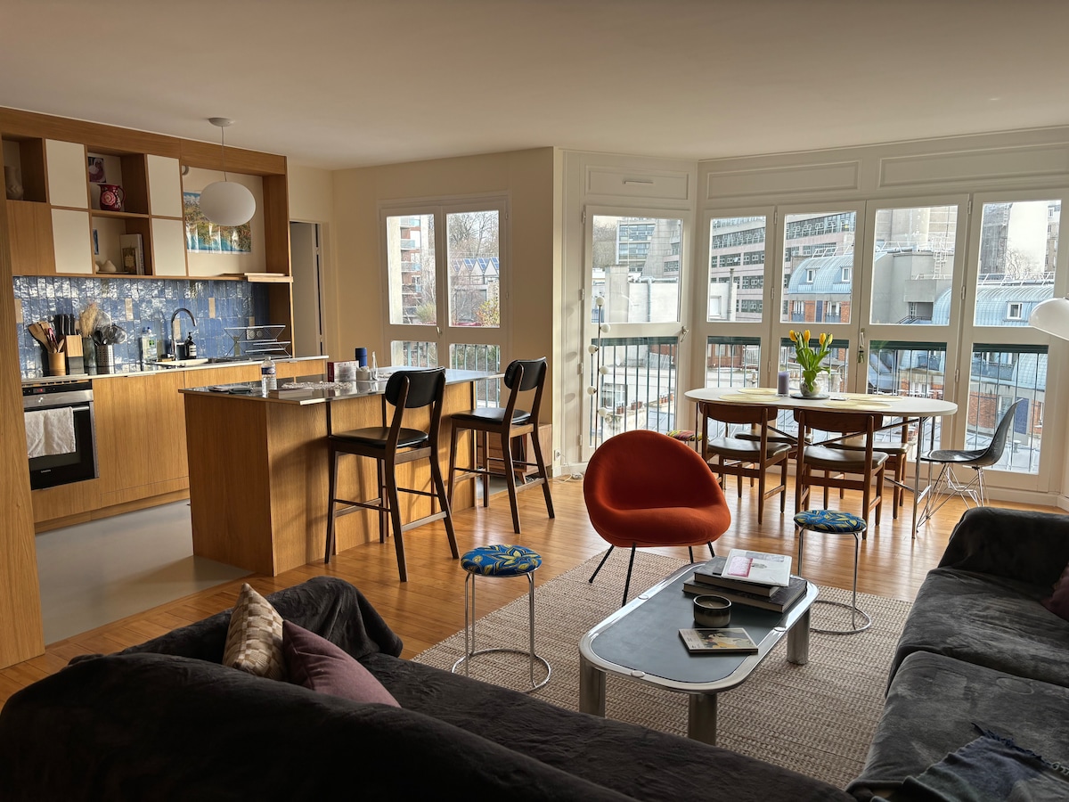 A spacious living area features large windows allowing abundant natural light, illuminating the modern kitchen and dining space. A comfortable couch faces a stylish coffee table, with colorful seating around the dining table. The open layout enhances the inviting atmosphere of the apartment.