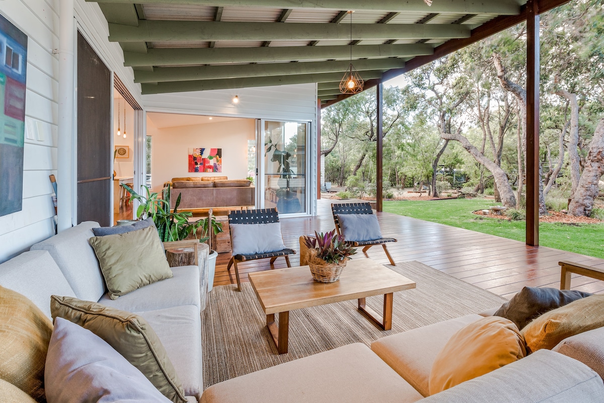 A covered outdoor seating area features comfortable sofas adorned with neutral and earthy-toned cushions. A wooden coffee table sits in the center, surrounded by greenery. Large glass doors lead to a spacious living area and the lush yard, enhancing the open feel of the space.