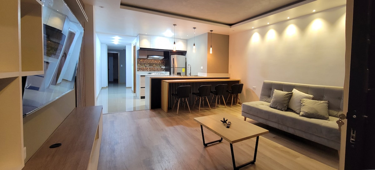 A modern living space features a spacious seating area with a gray sofa and a wooden coffee table. In the background, a well-equipped kitchen is visible, characterized by sleek cabinetry and pendant lighting. Soft lighting enhances the warm ambiance within the room.