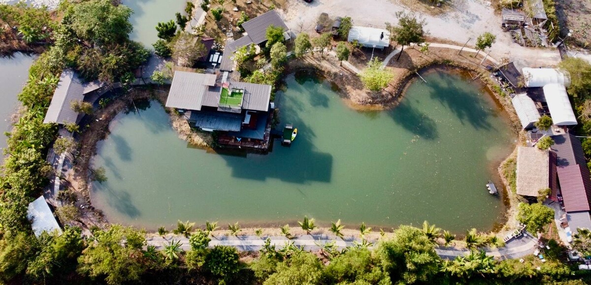 An aerial view reveals a serene landscape featuring a winding waterway bordered by lush greenery. Several structures, including a central house with a green roof, are situated along the water's edge, surrounded by trees and pathways.