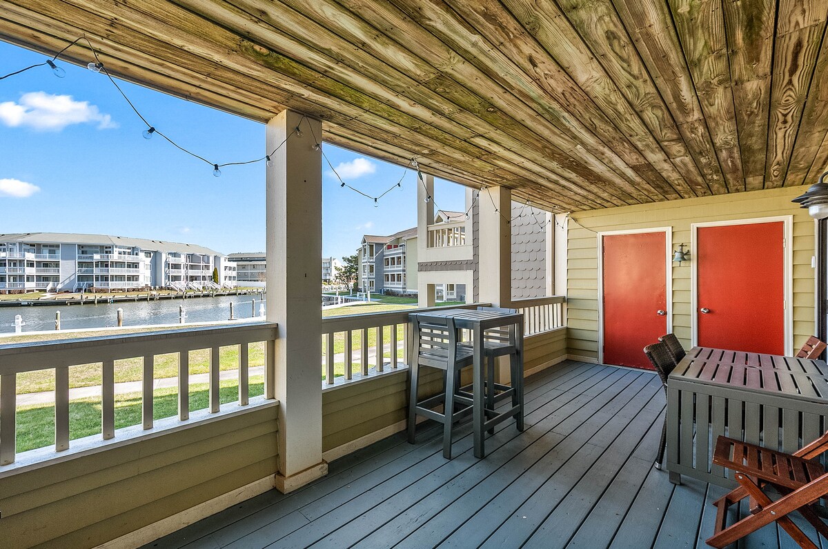 A spacious outdoor patio features a dining table and seating arrangements, surrounded by a wooden railing. The waterfront view showcases the canal and nearby buildings under a clear blue sky. String lights gently hang above, adding a touch of ambiance to the area.