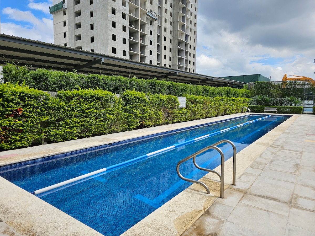 A long swimming pool features clear blue water, framed by light-colored tiled flooring. Surrounding greenery provides shade, while a metal railing is positioned at one end for safety. The pool area displays a relaxed atmosphere with ample space for lounging.
