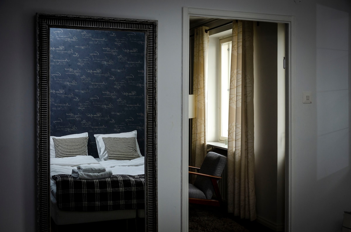 A spacious bedroom is visible through a large mirror, showcasing a neatly made bed with decorative pillows. The room features a dark wall with subtle design details. A doorway leads to a second area, where soft curtains frame a window and a comfortable chair is placed nearby.